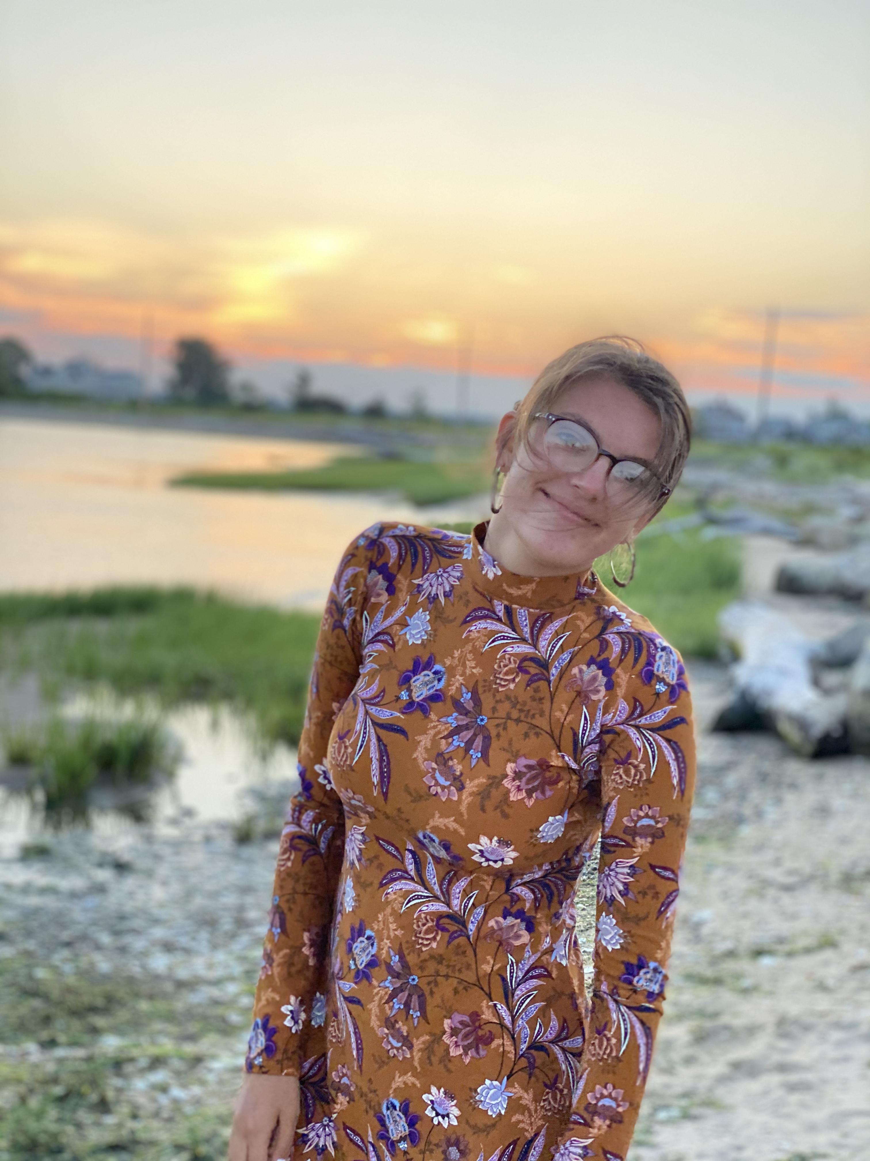 Paleena, wearing glasses and a brown and purple floral dress with a sunset behind her. 
