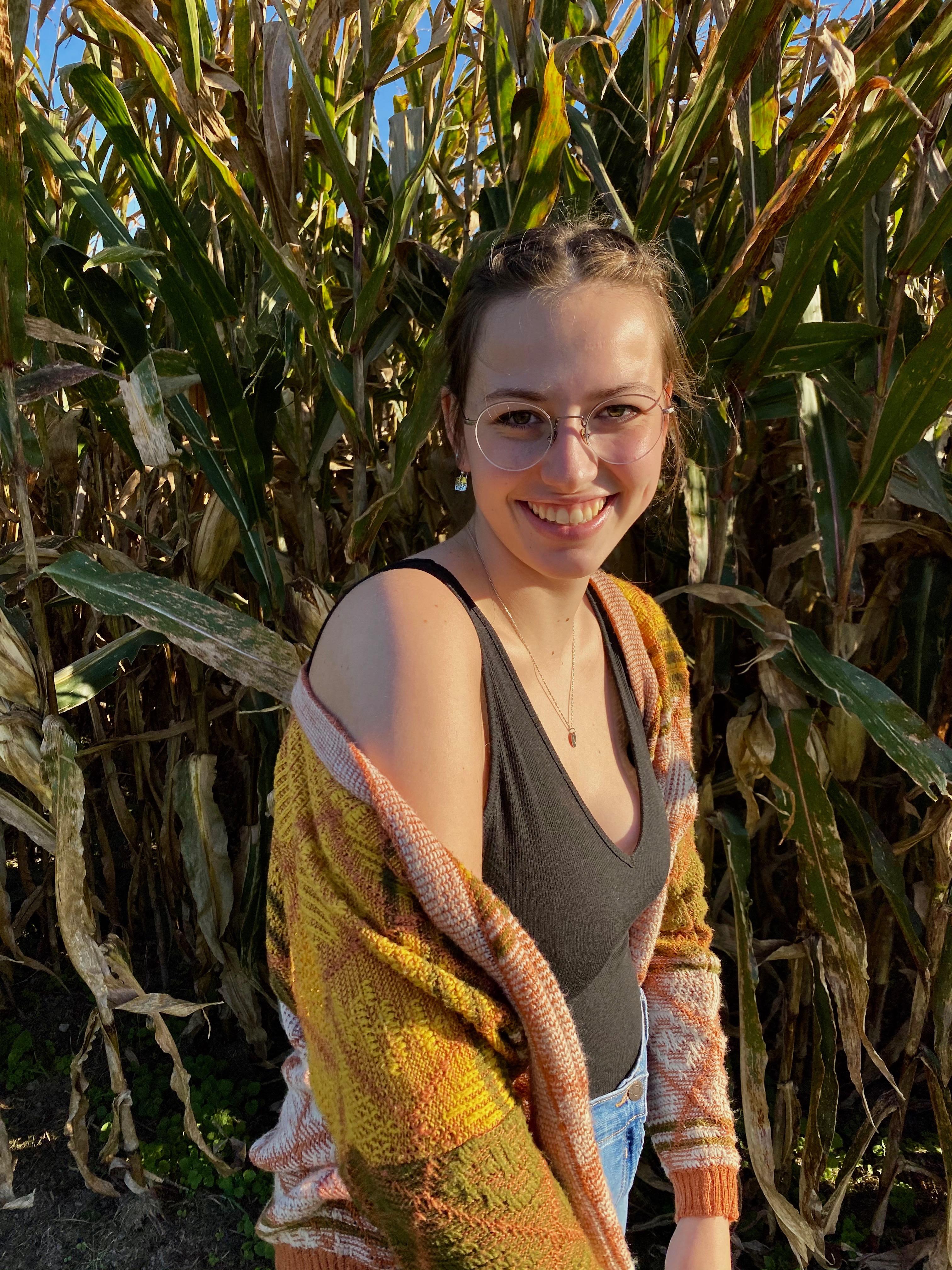 Sylvie, wearing a black shirt and multicolored cardigan, smiling with a field of corn behind her. 